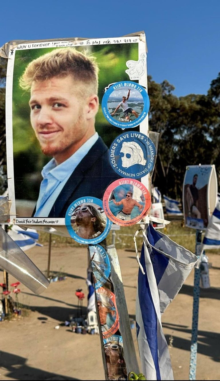 Memorial poster on wooden pole featuring portrait photo of young man with decorative badges and ribbons attached